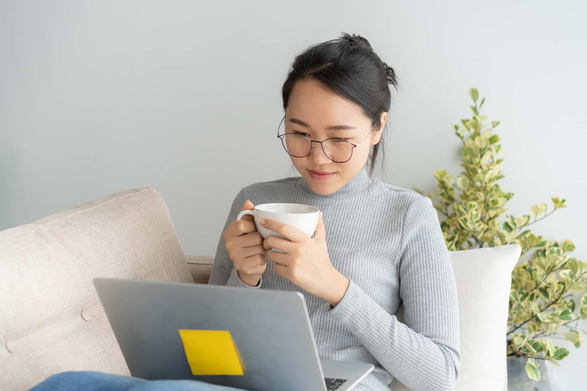 woman with glasses holding a cup reading on her laptop