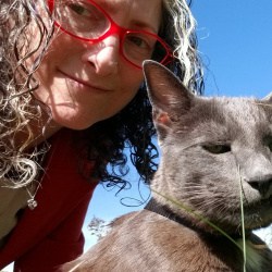 Woman with glasses and curly hair smiles at the camera next to her gray cat