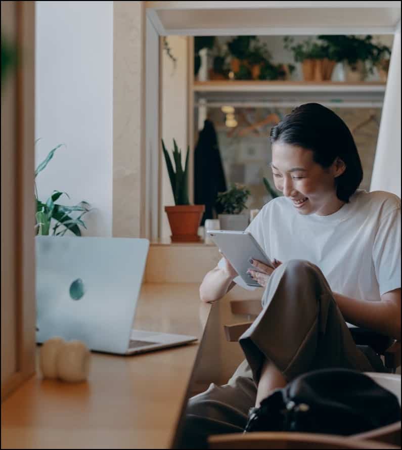 person with dark hair and brownish skin sits comfortably at a desk, reading a tablet and smiling