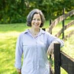 A white woman with gray hair cut in a bob smiles and leans against a fencepost