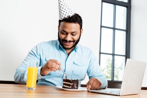 a man at a computer wearing a party hat eats a piece of cake